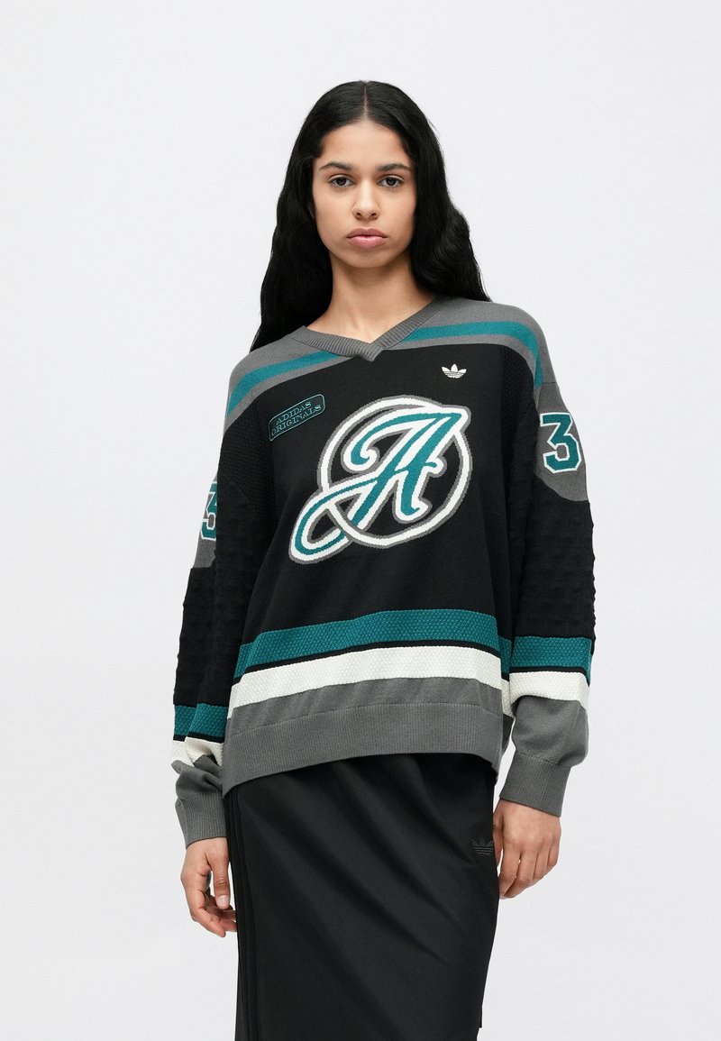 Pull en maille noire avec accents de rayures grises, bleu sarcelle et blanches ; il présente un col en V, une texture tissée et des détails brodés de logo et de numéro.