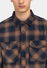 Marineblauwe en oranje geruite shirt van een zachte stof, met een puntkraag, twee borstzakken met knoopsluiting en een knopenlijst aan de voorkant.