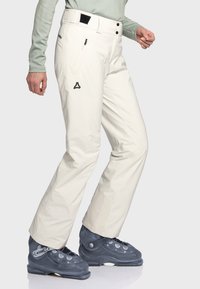 Damen Skihosen aus weißem Stoff mit glatter Textur, ausgestattet mit einem schwarzen Logo, einer Reißverschlusstasche und einer taillierten Passform über grauen Skischuhen.