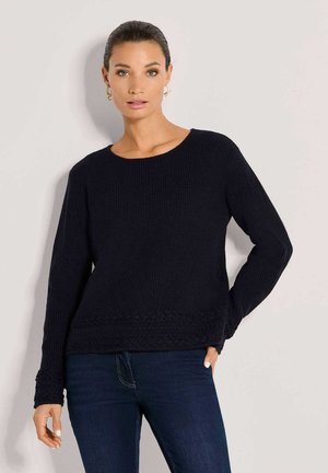 Vrouw die een zwarte gestructureerde lange mouw trui draagt met donkerblauwe spijkerbroek, staand tegen een lichte achtergrond.