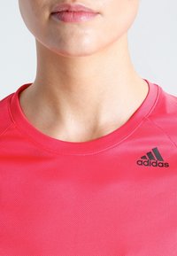 Maglietta sportiva rosa realizzata in tessuto leggero e texturizzato, con collo a giro. Presenta un logo Adidas nero sul lato sinistro.