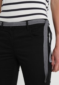 Svarta jeans med en figurnära stil, prydda med ett randigt svart och vitt bälte knutet vid midjan. Närbild visar fickor och sömmar.