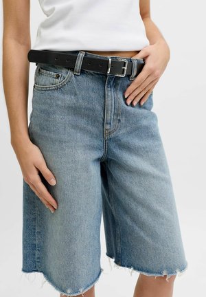 Person iført lyseblå denim Bermudashorts med rå kanter, en hvid ribbet tanktop og et sort læderbælte med sølvspænde.