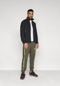 Μαύρο μπουφάν με φερμουάρ και πλευρικές πάνελ από δίχτυ, λευκό T-shirt, χακί πράσινο παντελόνι jogger με το λογότυπο "JORDAN", και μαύρα/κόκκινα αθλητικά παπούτσια.