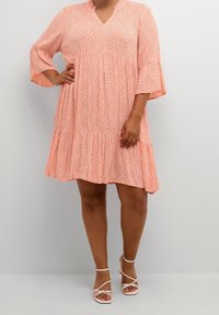 Robe florale rose clair avec un décolleté en V, des manches cloche trois quarts et une jupe à volants. Associée à des sandales à talons blanches à lanières.