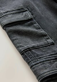 Tissu en denim noir avec une surface texturée. Présente une poche latérale avec rabat et des détails de couture, mettant en valeur un design décontracté et utilitaire.