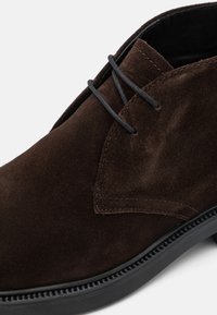 Brown suede shoe featuring a lace-up design, smooth texture, and a black rubber sole with decorative stitching.