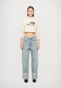 Skrátená béžová mikina s logom "Chino Jeans", v kombinácii s svetlomodrými širokými nohavicami so pruhovanými manžetami. Čierna obuv dotvára celý outfit.