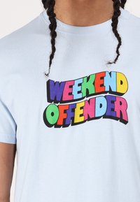 Šviesiai mėlynas medvilnės marškinėliai su ryškiu, daugiaskaitiniu tekstu, rašančiu "WEEKEND OFFENDER" įvairiomis formomis ir dydžiais.