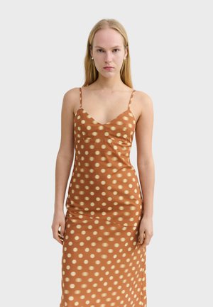 Woman with long blonde hair wearing a brown slip dress with cream polka dots, standing against a plain light background.
