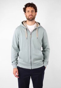 Veste à capuche zippée bleu clair en tissu doux, avec une doublure beige, un capuchon réglable et des poches avant. Associée à un pantalon foncé.