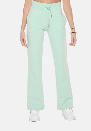 Femme portant un pantalon vert clair à cordon et des baskets blanches, debout les mains relâchées le long du corps devant un fond uni.