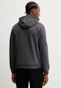 Felpa grigia con cappuccio e vestibilità ampia, caratterizzata da un particolare dettaglio sul retro. Il look è completato da un tessuto morbido e polsini a costine.