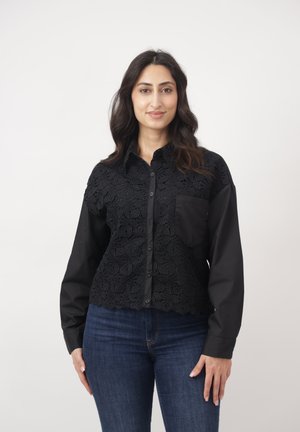 Kobieta o długich ciemnych włosach, ubrana w czarną koszulę na guziki z koronkowym wzorem oraz niebieskie dżinsy, stojąca na tle jednolitego jasnego tła.