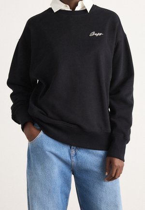 Personne portant un sweat-shirt noir avec un petit logo blanc en écriture manuscrite sur une chemise blanche à col et un jean bleu clair, main dans la poche.