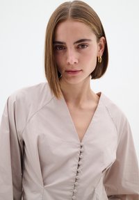 Abito beige chiaro con scollo a V, dettagli di bottoni lungo la parte frontale e maniche lunghe. Tessuto liscio e testurizzato con una silhouette aderente.