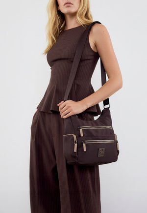 Bolso de hombro de nailon marrón con dos bolsillos frontales con cremallera, acentos en contraste color crudo y un parche con logo. El bolso cuenta con una correa larga y ajustable.