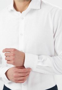 Bílá košile s dlouhým rukávem, klasickým límcem, zapínáním na knoflíky vpředu a detaily na manžetách, vyrobená z hladké látky bez viditelných vzorů nebo akcentů.