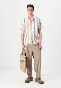 Camisa de manga curta com riscas verticais em cores pastel (laranja, verde, azul), combinada com calças largas bege e sapatos a condizer.