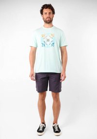 Camisa de color azul claro con una estampado gráfico en naranja y azul verdoso, combinada con pantalones cortos de carga oscuros y zapatillas negras con cordones blancos.