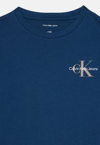 Mėlynas medvilninis marškinėliai su apvalia iškirpte. Kairėje krūtinės pusėje yra spauda "Calvin Klein Jeans" ir stilizuotas "CK" logotipas šviesiai smėlio spalvos.