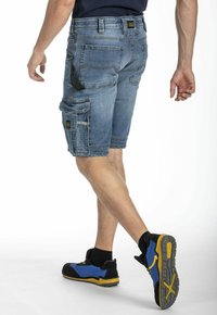 Shorts cargo en denim bleu clair, avec une grande poche latérale, des détails cousus et une coupe fuselée. Associés à des chaussures noires et bleues.
