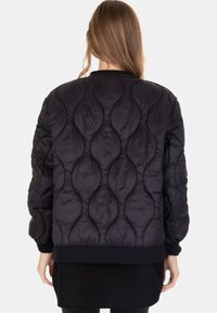 Donna con lunghi capelli castani che indossa una giacca imbottita nera con motivi imbottiti circolari, vista di spalle su uno sfondo bianco.