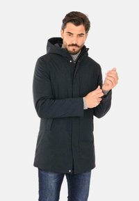 Parka nera con cappuccio, dotata di zip frontale e chiusura a scatto. Caratterizzata da tasche laterali e una texture liscia. Indossata sopra un maglione di maglia grigia.
