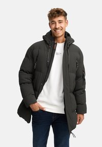Polstret mørkegrøn jakke med hætte, tekstureret stof, frontlynlås og flere lynlåslommer. Båret over en hvid sweatshirt og blå jeans.