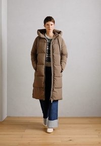 Doudoune taupe avec capuche, fermeture éclair à l'avant et design matelassé. Portée sur un pull à motifs et un jean en denim évasé avec des ourlets retroussés.