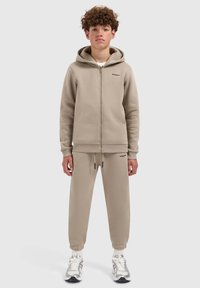 Beige zip-up hoodie met een kangoeroezak en bijpassende tapered joggers. Glad materiaal, geribbelde manchetten en logo-detail op beide stukken.