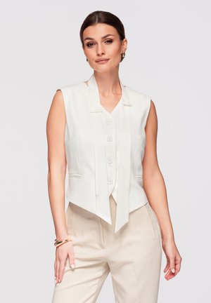 Donna con capelli castani raccolti in uno chignon basso, che indossa un gilet bianco senza maniche con bottoni sul davanti e pantaloni beige a vita alta, posando davanti a uno sfondo semplice.