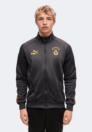 Jeune homme aux cheveux courts et bouclés portant une veste noire zippée avec le logo Puma doré et l'emblème du club de football Galatasaray.