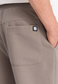 Beige ribbed tkaninové kalhoty se zadní kapsou, na které je černobílé logo. Textura je měkká a vzorovaná vodorovnými pruhy.