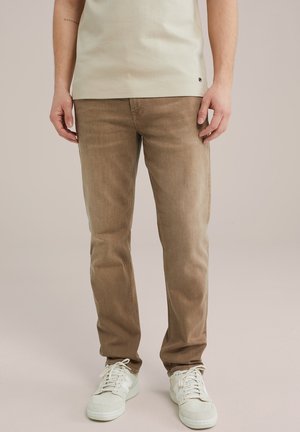 Man die lichtbruine spijkerbroek, witte sneakers en een beige overhemd draagt, staand tegen een effen achtergrond.