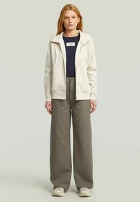 Woman standing wearing cream zip-up hoodie, dark navy logo t-shirt, wide-leg grey pants, and beige sneakers against plain background.