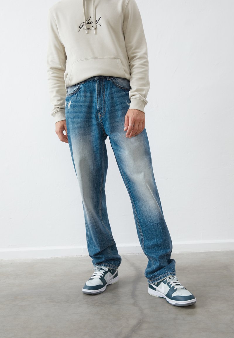 Light grey hoodie with front pocket and logo, paired with blue straight-fit jeans featuring fading. Grey and white sneakers complete the look.