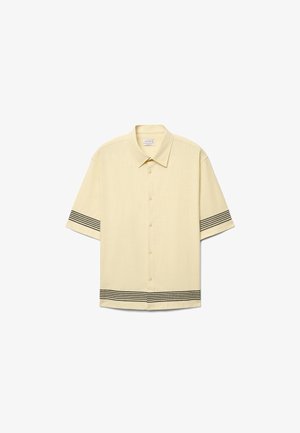 Beiges Kurzarmhemd mit Knopfleiste, schwarzen horizontalen Streifen an den Ärmeln und am unteren Saum, mit spitzem Kragen.