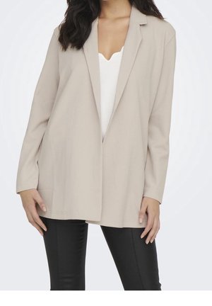 Donna che indossa un lungo blazer beige sopra una maglietta bianca e pantaloni neri, in piedi con le braccia rilassate lungo i fianchi contro uno sfondo chiaro.