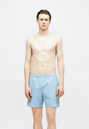 Jeune homme aux cheveux courts et foncés, debout, torse nu et portant un short de bain bleu clair sur un fond blanc uni.