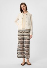 Kremowa kurtka z dwiema przednimi kieszeniami, zestawiona z szerokimi spodniami w zygzakowy wzór w odcieniach beżu, brązu i czerni. Brązowe sandały.