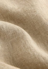 Tissu beige tissé avec une texture grossière, montrant de petites bosses et plis créant des ombres douces et de la profondeur.