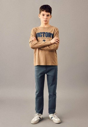 Brown long-sleeve shirt with blue text "VICTORY" worn with dark blue jeans and white sneakers. The outfit features a relaxed fit.