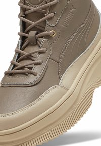 Zapatilla alta de cuero marrón con suela de goma beige, con costuras en crema, ojales dorados y diseño de cordones.