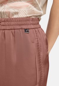 Rosa Satinhose mit elastischem Bund, seitlichen Nähten und einem kleinen Markenetikett. Glatte Textur und lockere Passform.