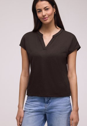 Junge Frau mit langem dunklem Haar, die goldene Creolen, ein dunkelbraunes kurzärmeliges V-Ausschnitt-Top und hellblaue Jeans vor einem neutralen Hintergrund trägt.