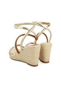 Sandalias de cuña con tacones de paja trenzada, correas metálicas en oro y marrón, y cierres de hebilla en el tobillo, mostradas desde un ángulo trasero.