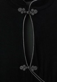 Prenda negra con escote curvado, decorada con cierres en forma de nudos en espiral grises y un acabado en contraste a lo largo de la apertura.