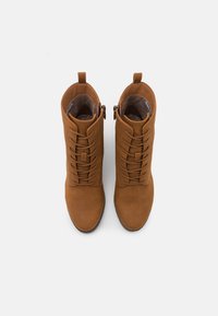 Bottines en suède marron avec un laçage à l'avant, texture lisse et fermeture éclaire sur le côté. Elles présentent une pointe arrondie et une languette à l'arrière.