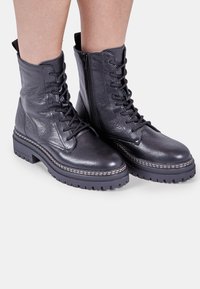 Botine negre din piele cu un finisaj texturat, cu sireturi în față, fermoar lateral și talpă groasă din cauciuc accentuată cu cusături albe.